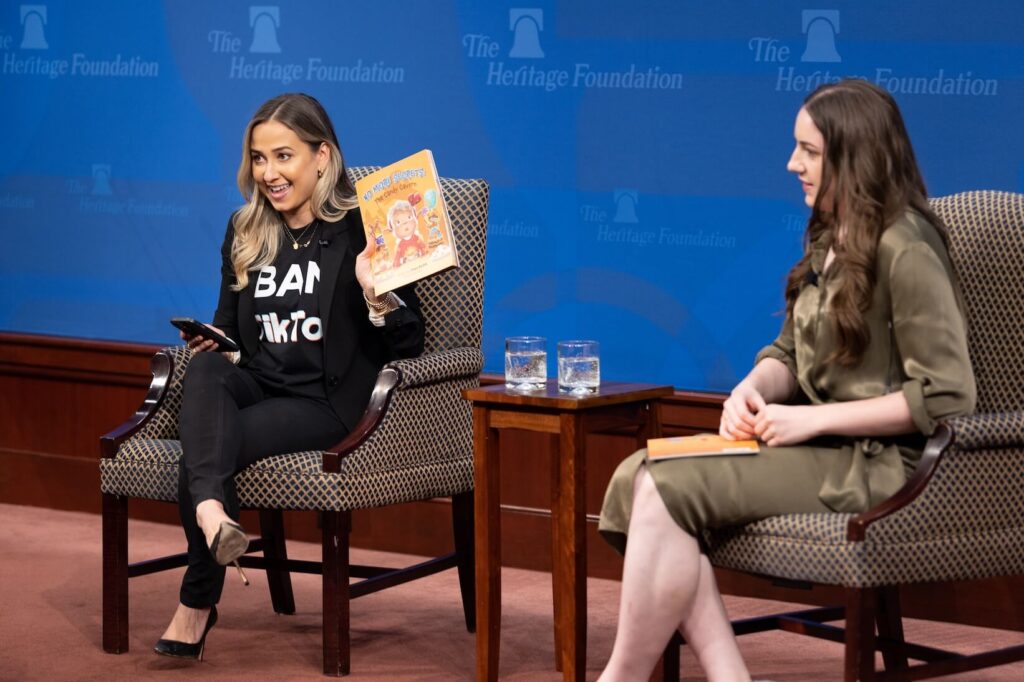 The Heritage Foundation's Kara Frederick, Director of Tech Policy, at an event wearing a "Ban TikTok" shirt and holding a book on stage, seated next to another woman speaker