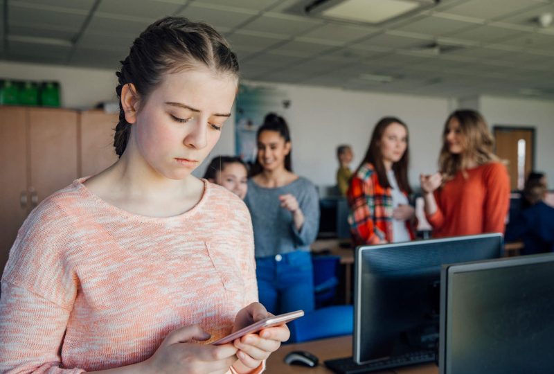 girl looking at phone getting bullied and students in background laughing and pointing at her